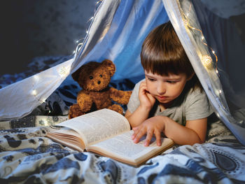 Little boy reads book. toddler plays in tent made of linen sheet with light bulbs on bed. 