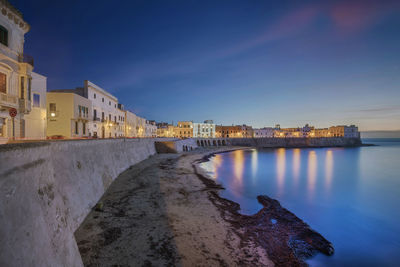 Illuminated buildings by sea against sky at sunset