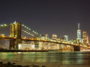 Illuminated bridge over river by buildings against sky at night