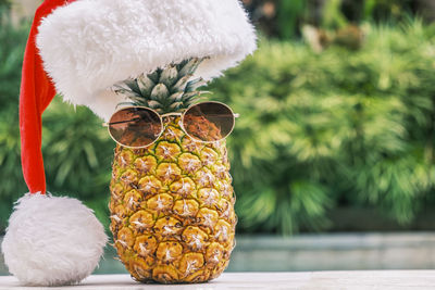 Funny pineapple wearing sunglasses and santa claus hat against pool and tropical plants 