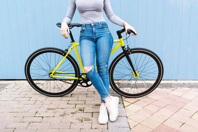 Mid adult woman wearing torn jeans sitting on fixie bike against blue wall