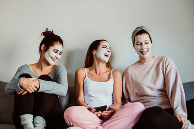 Cheerful female friends with facial masks sitting against wall at home