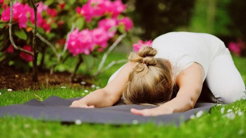 A blond woman wearing white yoga suit practicing yoga in green park between blooming flowers