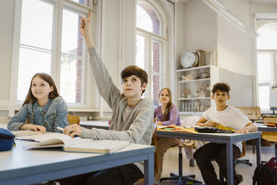 Boy raising hand while sitting with female friend at desk in classroom
