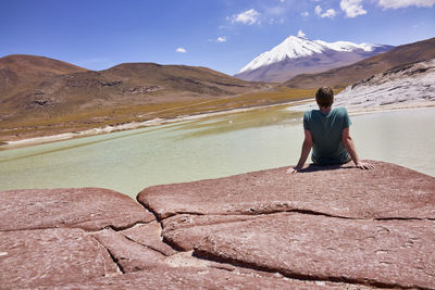 Rear view of man sitting on rock at lake against sky