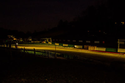 Illuminated street by field against sky at night