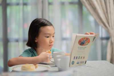 Girl eating food while reading book on table at home