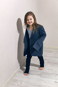 Funny portrait of fashionable girl child in large gray jacket stands against gray wall next  window 