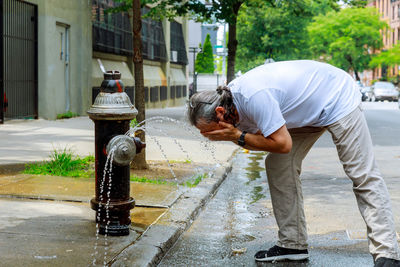 Side view of man washing face in flowing water from fire hydrant in city
