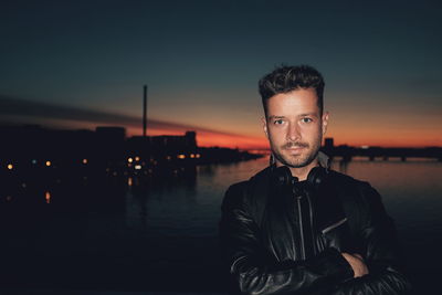 Portrait of young man with arms crossed standing by sea against sky during sunset