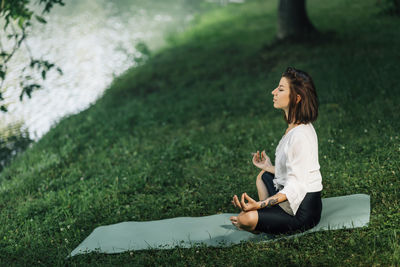 Young woman sitting in lotus position and practicing meditation near water in the nature