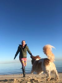 Woman running towards dog at beach against clear blue sky