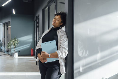 Young businesswoman with eyes closed leaning on glass in office