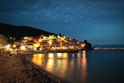 Illuminated buildings by sea against sky at night