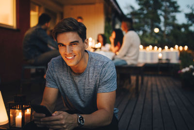 Portrait of smiling young man holding smart phone while friends in background during dinner party