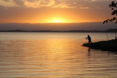 Silhouette man fishing by sea against sky during sunset