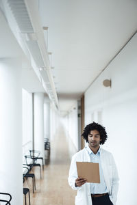 Confident young male doctor looking at clipboard in hospital corridor