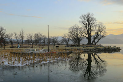 Bare trees by lake against sky during sunset