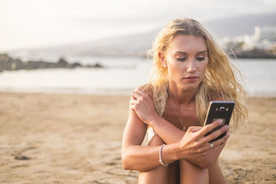 Young woman using mobile phone while sitting at beach against sky