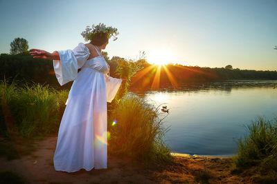 Rear view of woman standing against lake during sunset