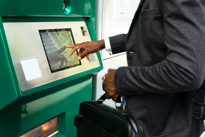 Cropped side view of unrecognizable african american businessman in protective mask with suitcase using street atm terminal in barcelona city