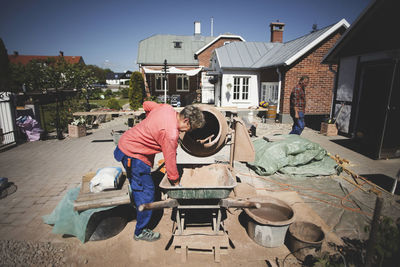 Senior woman using cement mixer at yard while man walking in background