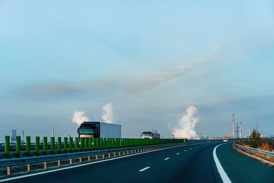 Trucks on highway by factory with steam against morning sky during autumn