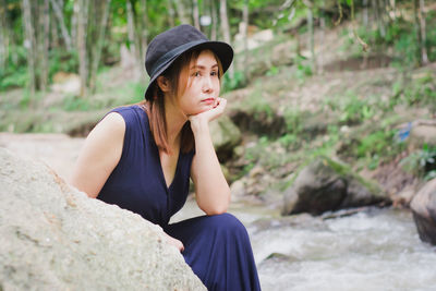 Woman with hand on chin looking away while sitting on rock by river