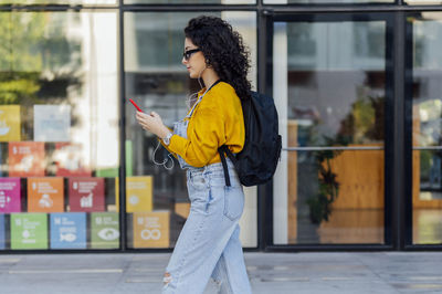 Woman listening to music through in-ear headphones and using smart phone in front of glass building