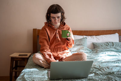 Teen girl watching series movie on laptop sitting in bed drinking tea at home.