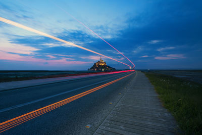 Light trails on road by field against cloudy sky at dusk