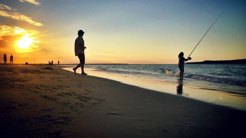 People on shore at beach against sky during sunset