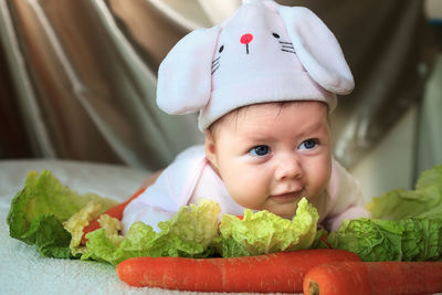 Close-up of cute baby girl wearing bunny hat while lying with carrots and lettuce on bed