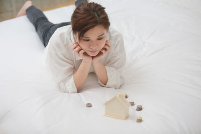 High angle view of looking at model house with coins while lying on bed at home