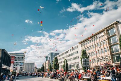 Low angle view of balloons flying over crowd and street amidst buildings on sunny day