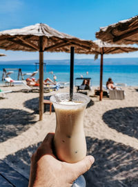 Personal perspective of person holding glass of cold iced coffee on beach in summer.