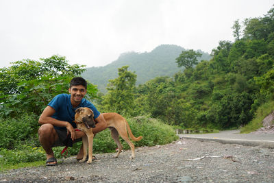 Young man sitting with his dog beside a road with blurred mountains in the background.