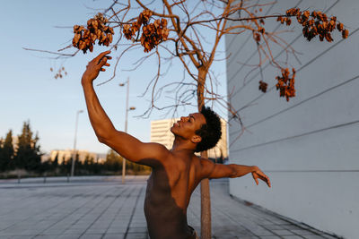 Shirtless man dancing against bare tree in city