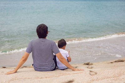 Father and son are sitting on the beach by the sea in summer in white t-shirts with their rear