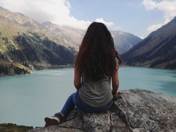 Rear view of woman sitting on rock by lake against sky