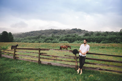 Young man standing by fence on grassy field against sky