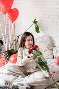 Young caucasian brunette woman sitting in the bed celebrating valentine day smelling red roses