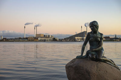 Statue by river against sky during sunset