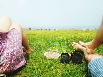 Low section of couple resting by champagne flutes on grassy field against clear sky