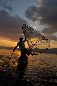 Fisherman holding fishing cage while standing on boat in inle lake against sky during sunset