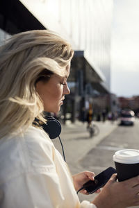 Side view of woman using mobile phone while holding coffee cup in city