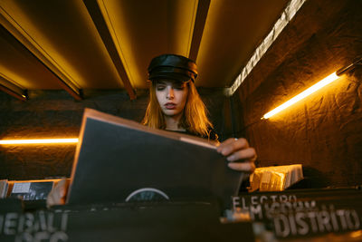 Front view of serene young female in black cap choosing vinyl record while standing near counter with collection of musical albums in store with glowing lamps