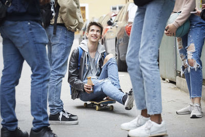 Low section of friends standing by smiling teenage boy sitting on skateboard