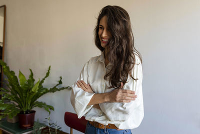 Smiling female model with arms crossed standing against wall at home