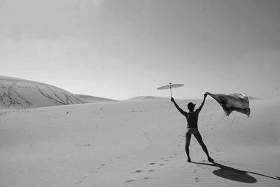 Rear view of man standing on sand covered landscape against sky with umbrella and flag